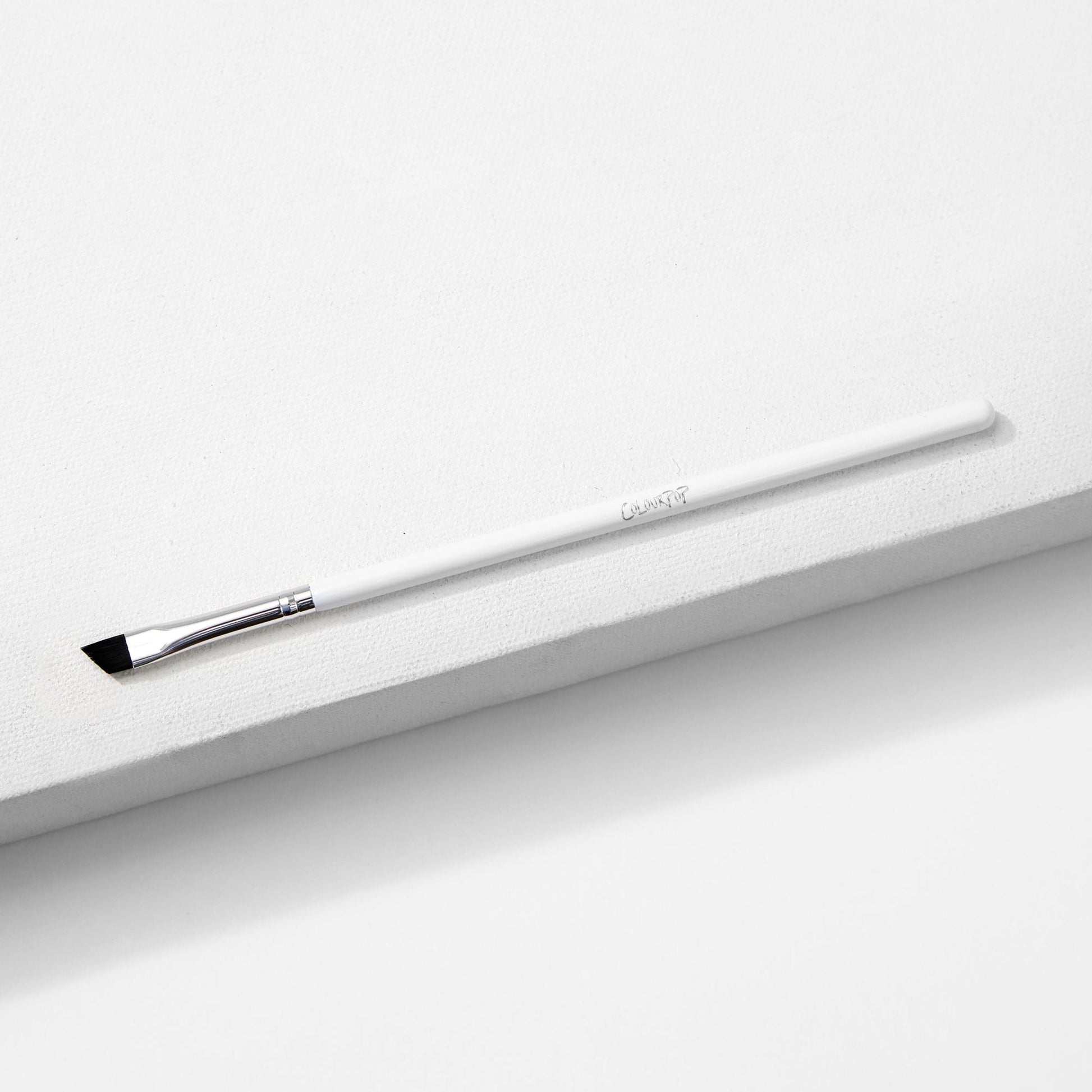 ColourPop Angled Eye Brush resting diagonally on a white textured block, featuring slim angled black bristles, a silver ferrule, and a long white handle - makeup brush for precise eyeliner and brow shaping.