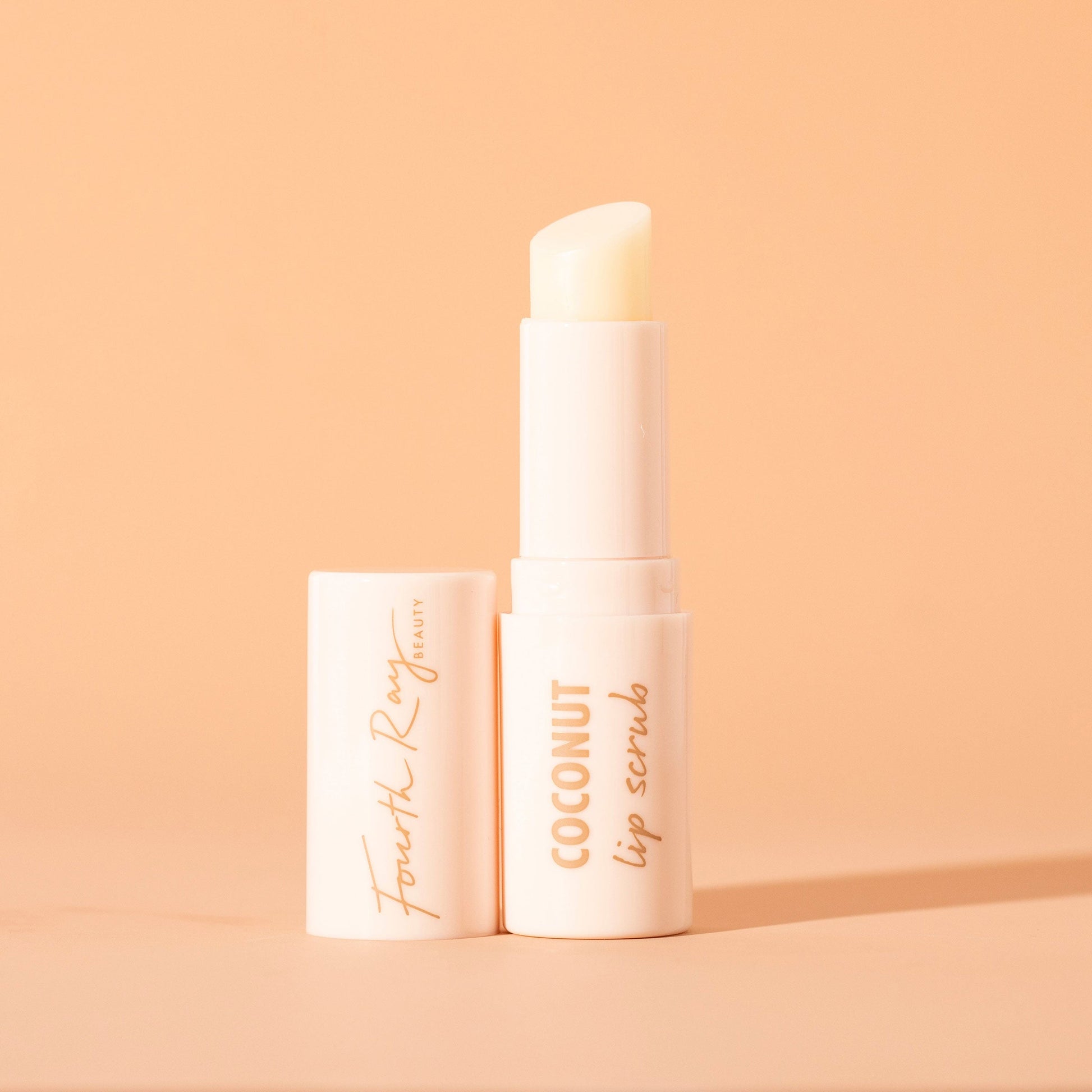 Coconut ColourPop Lip Scrub stick stands upright with the cap off, revealing an angled balm in a sleek ivory Fourth Ray Beauty tube with gold “COCONUT lip scrub” text on a soft peach background.