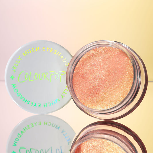 Open pot of ColourPop Jelly Much Shadow in Buzzy, a peachy coral-gold shimmer, sitting beside a white lid with iridescent lettering on a mirrored surface against a warm gradient backdrop.