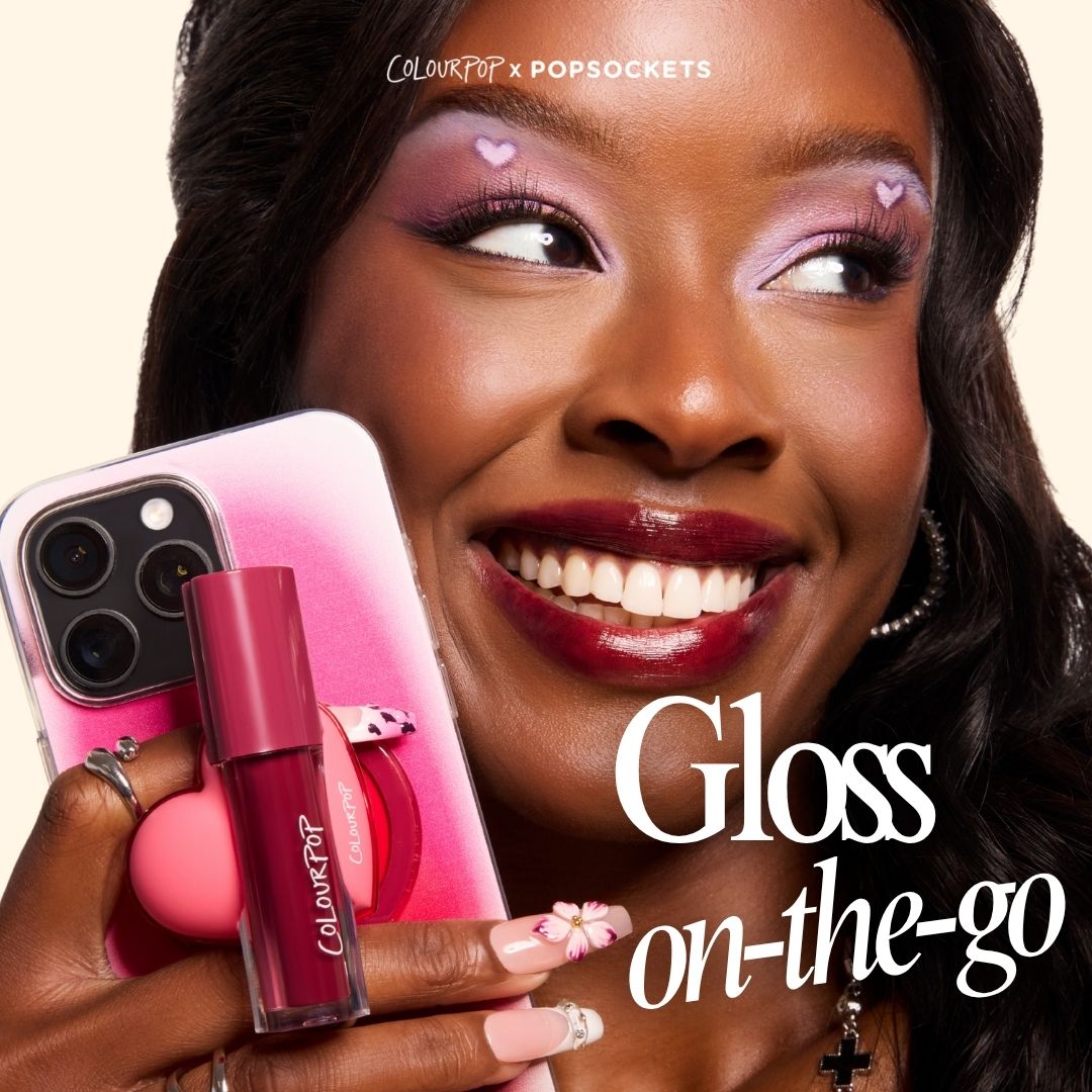 ColourPop Lip Stain tube in deep berry held against a pink smartphone with a ColourPop PopSocket, as a smiling model shows glossy stained lips and lilac heart eye makeup; ColourPopSockets collab highlights on-the-go lip color.